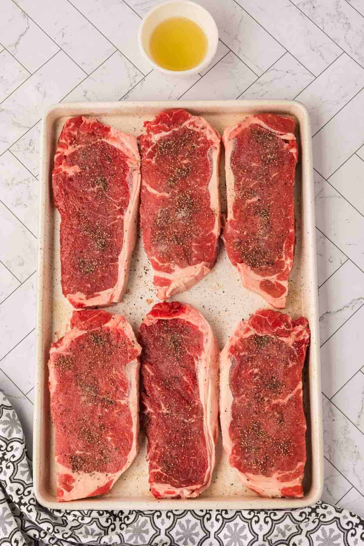 Six raw, seasoned Garlic Butter Steaks are arranged on a baking sheet with a small bowl of oil above them. The background features a patterned white countertop and a Blackstone-themed black-and-white decorative cloth in the corner.