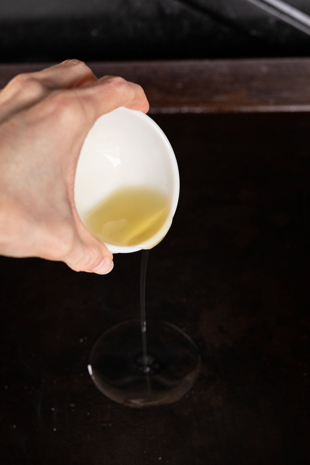 A hand pours a small amount of oil from a white cup onto a dark Blackstone Grill, preparing the surface for delicious Garlic Butter Steak.