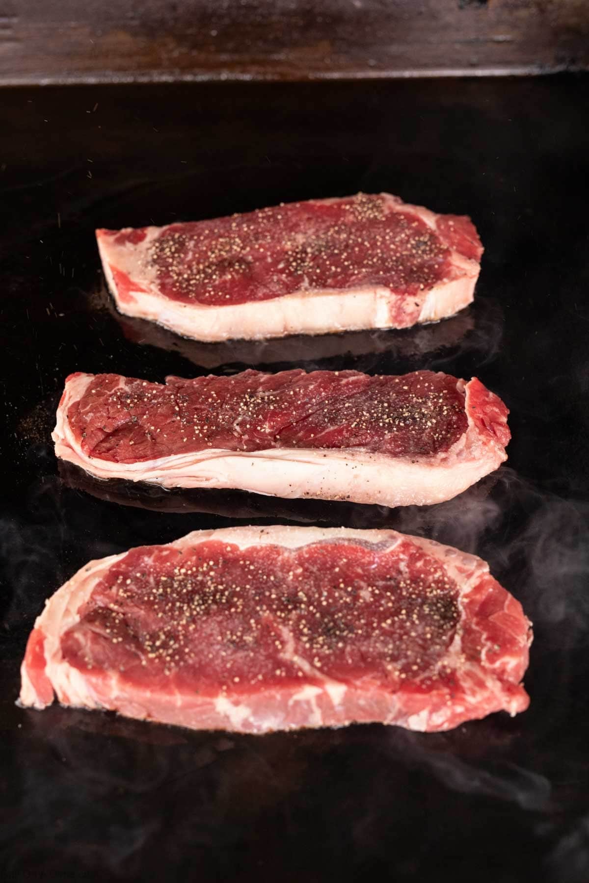 Three raw, seasoned steaks with visible fat marbling sizzle on a hot Blackstone griddle, steam rising as they cook to perfection—ideal for crafting a mouthwatering Garlic Butter Steak.