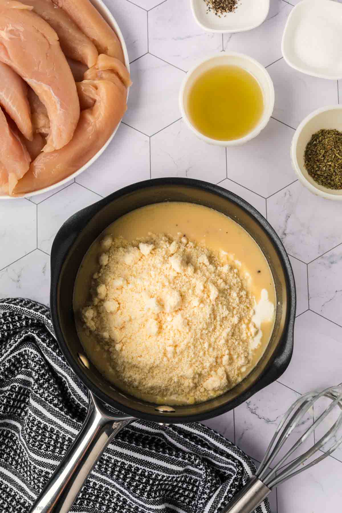 A saucepan filled with a creamy mixture and grated cheese sits beside a bowl of raw Garlic Parmesan Chicken strips, olive oil, spices, and a black-and-white towel on a white hexagon-patterned surface.