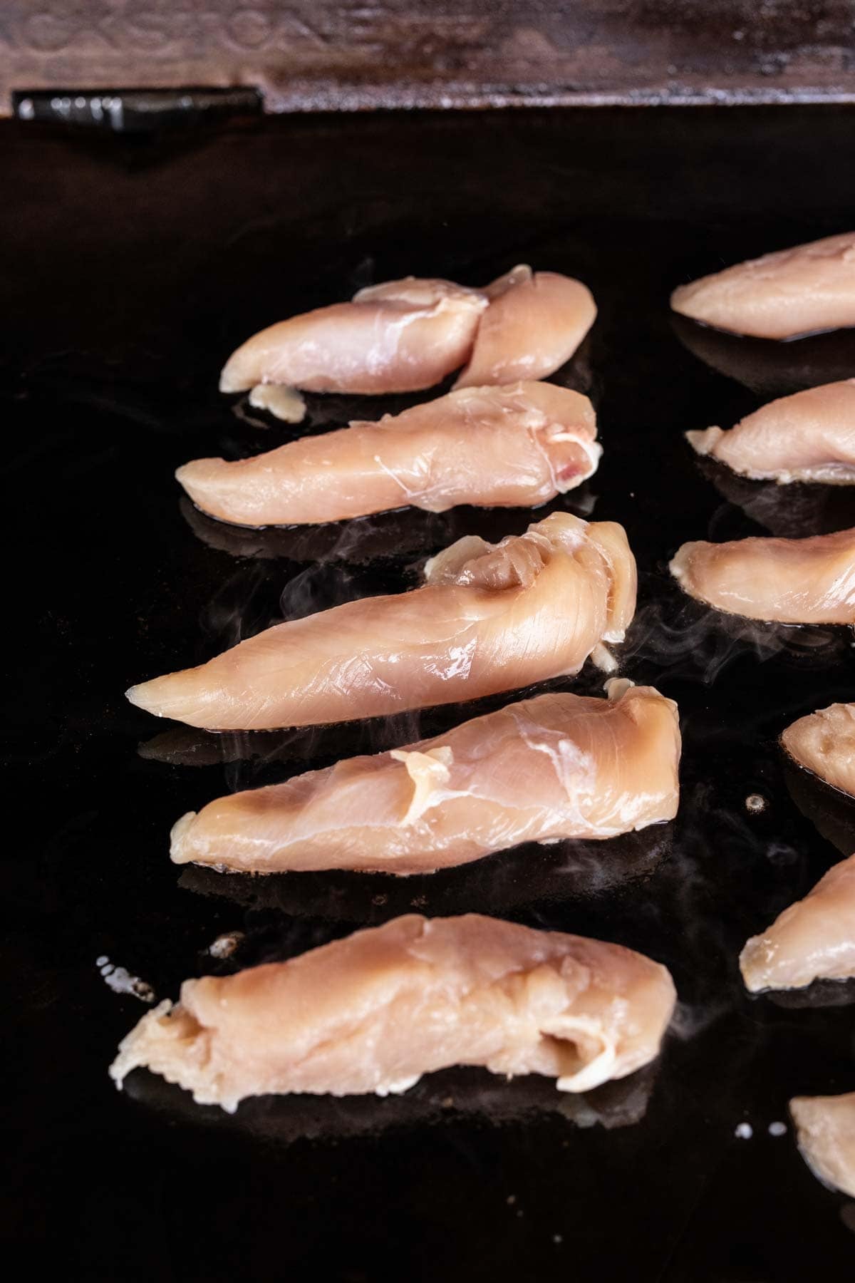 Raw chicken tenders arranged in rows on a Blackstone griddle, ready to be cooked into delicious Garlic Parmesan Chicken. Steam is visible rising from the meat.