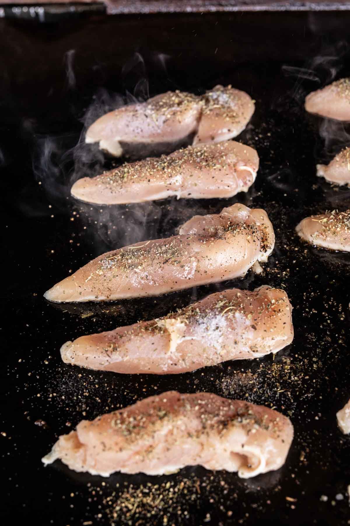 Raw chicken breast strips seasoned with spices are sizzling on a hot Blackstone griddle, with steam rising as they begin to cook—perfect for creating savory Garlic Parmesan Chicken.