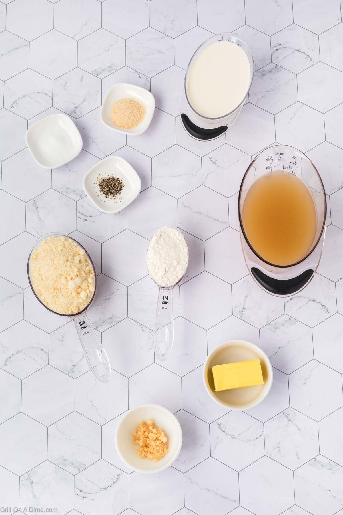 Ingredients for a Garlic Parmesan Chicken recipe arranged on a white hexagon-tiled surface, including grated cheese, flour, butter, minced garlic, milk, broth, and small bowls of seasonings like salt, pepper, and garlic powder.