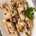 Grilled Garlic Parmesan Chicken breasts with herbs and a creamy white sauce, garnished with chopped parsley, served on a white platter with a sprig of fresh parsley—a delicious chicken recipe perfect for your Blackstone grill.