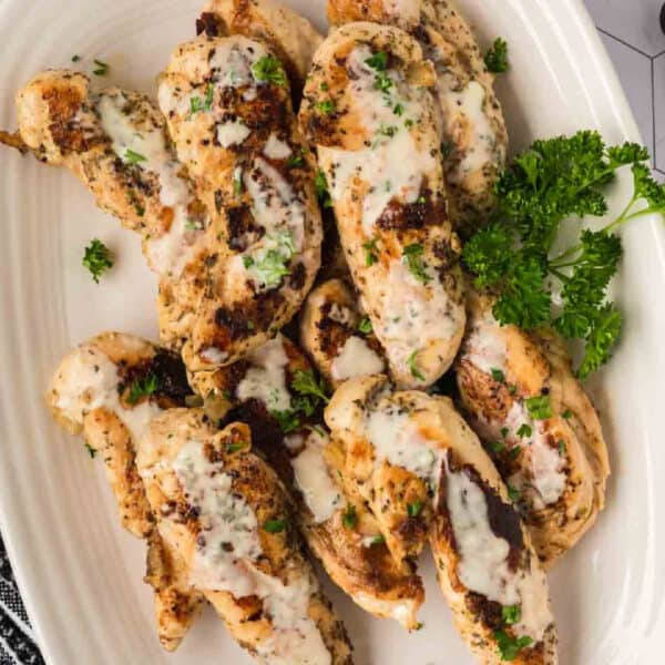Grilled Garlic Parmesan Chicken breasts with herbs and a creamy white sauce, garnished with chopped parsley, served on a white platter with a sprig of fresh parsley—a delicious chicken recipe perfect for your Blackstone grill.