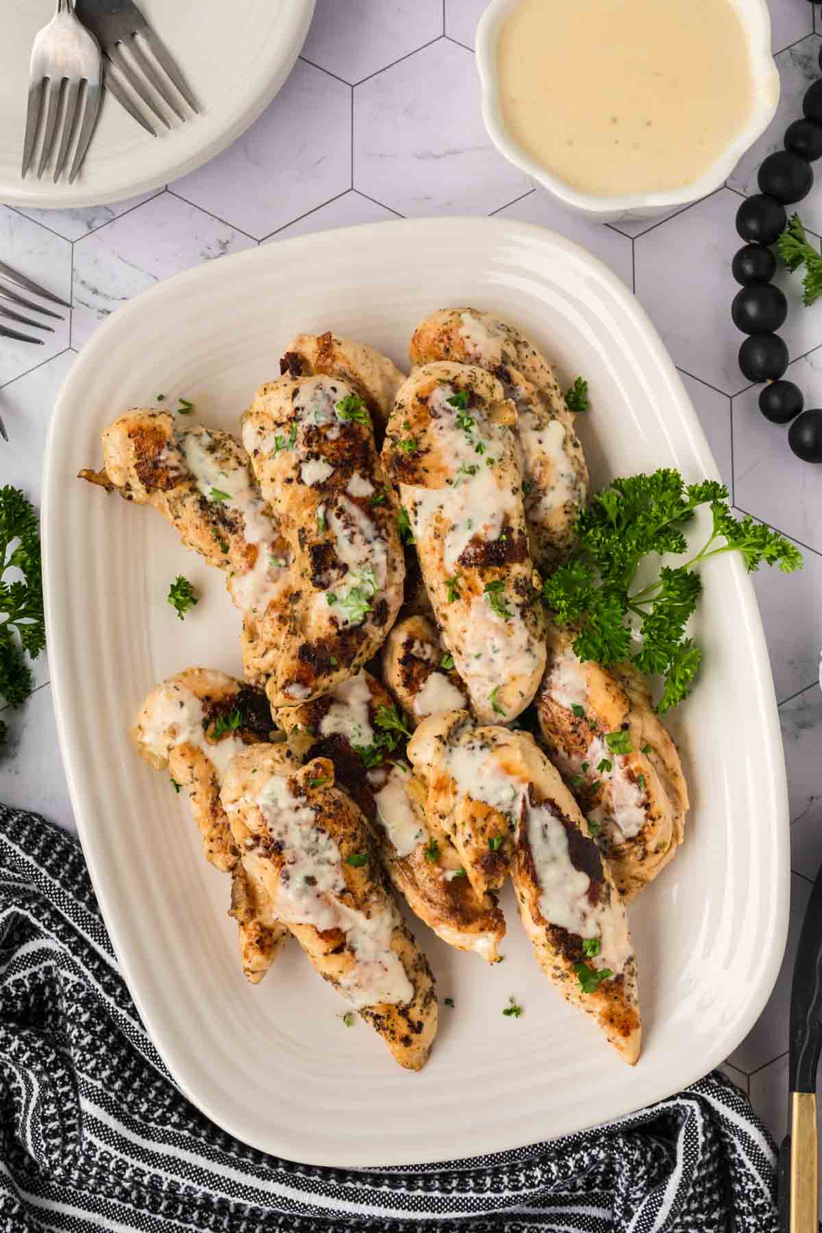 Grilled Blackstone Chicken breasts served on a white platter, topped with creamy garlic Parmesan sauce and garnished with parsley. A bowl of extra sauce, fork, knife, and napkin are nearby on a light patterned table.