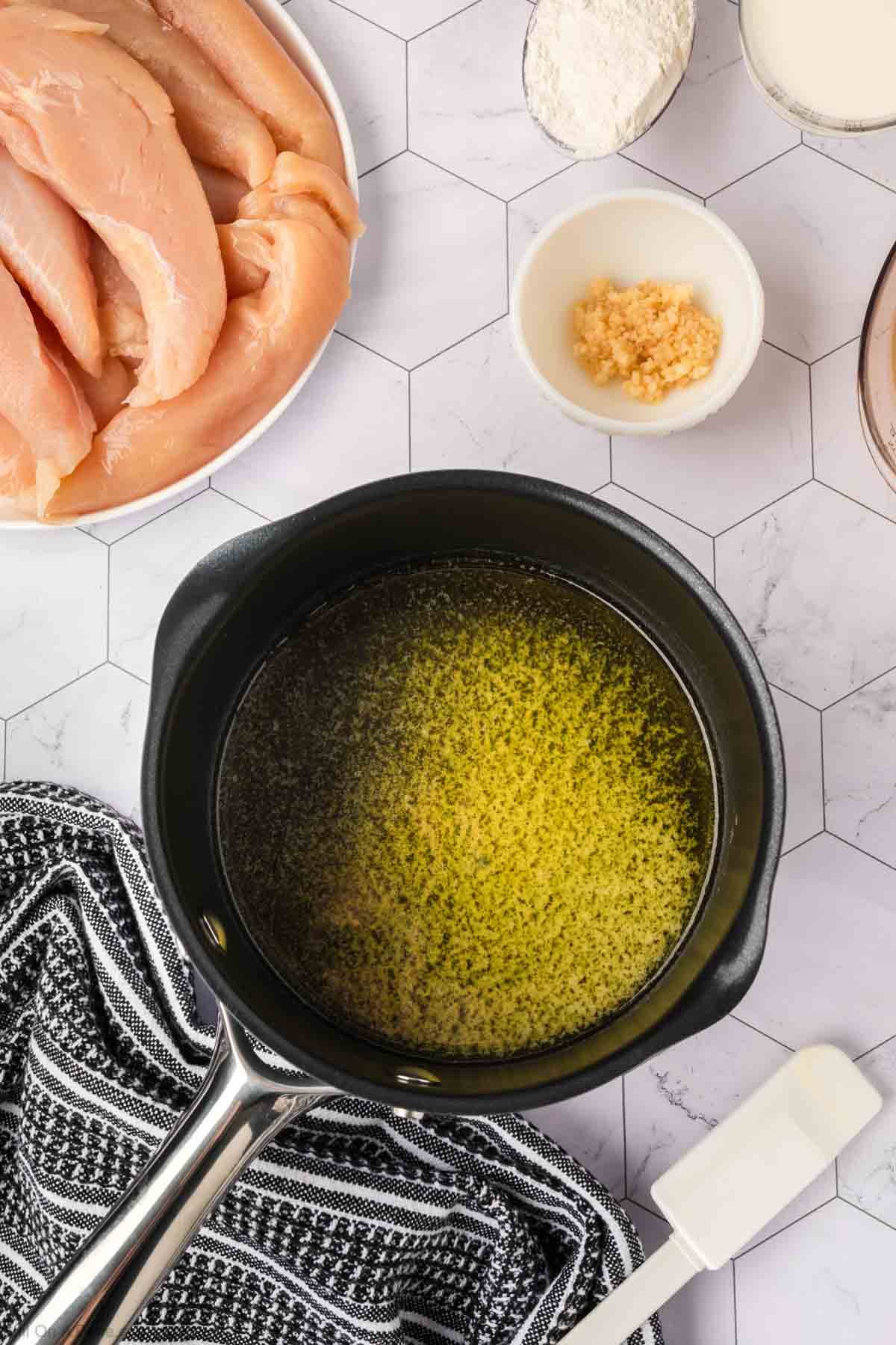 A saucepan filled with melted butter sits on a hexagon-tiled counter, surrounded by raw chicken strips, flour, and minced garlic in small bowls—ingredients for Garlic Parmesan Chicken—alongside a black-and-white kitchen towel.