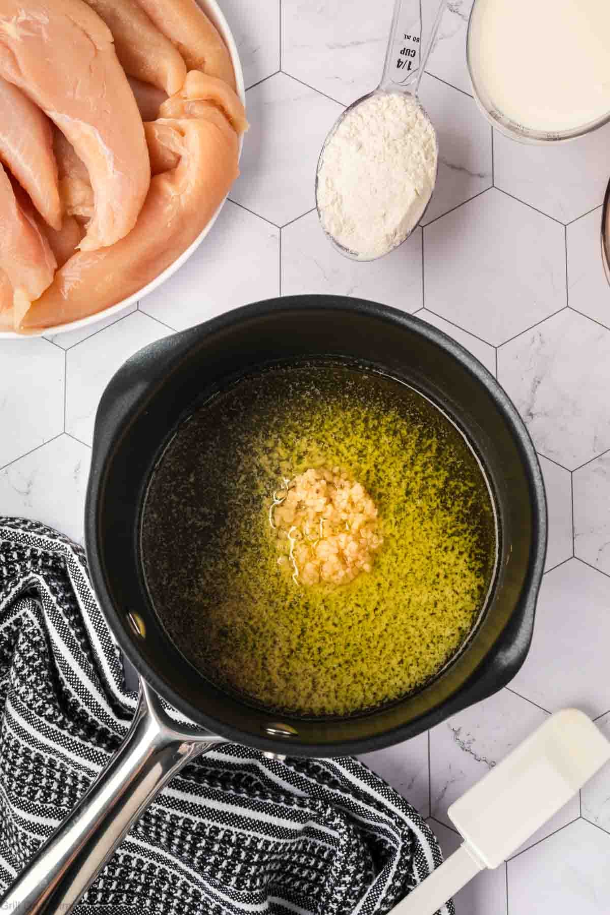 A saucepan with melted butter and minced garlic sits on a countertop beside ingredients for Garlic Parmesan Chicken: raw chicken breast, a measuring cup of flour, milk, and a black-and-white striped kitchen towel—perfect for your next Blackstone cookout.