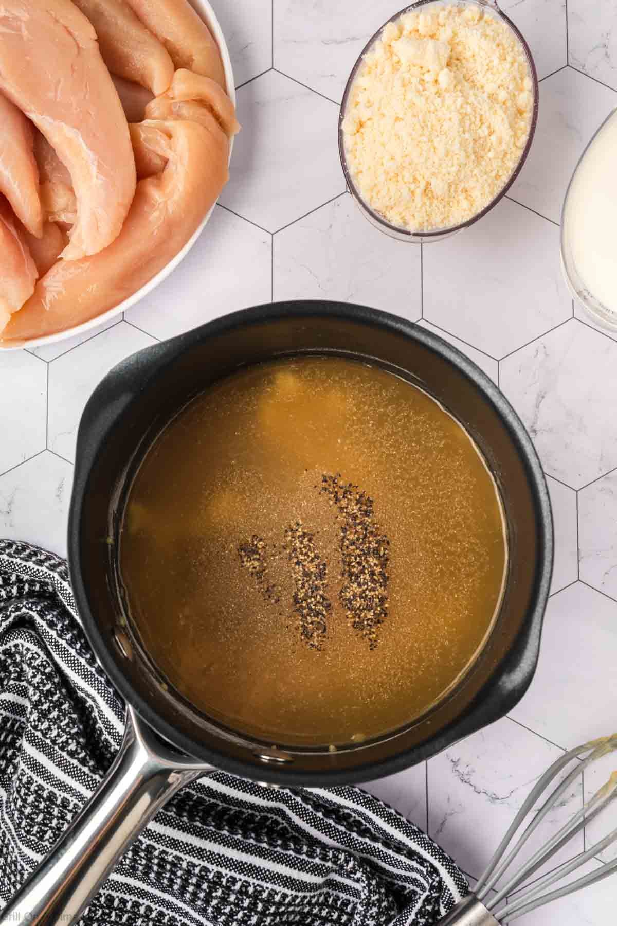 A saucepan of broth and black pepper sits on a striped towel. Nearby, bowls of raw chicken, grated cheese—perfect for Garlic Parmesan Chicken—and a glass of milk rest on a hexagonal-tiled surface, ready for the Blackstone grill.
