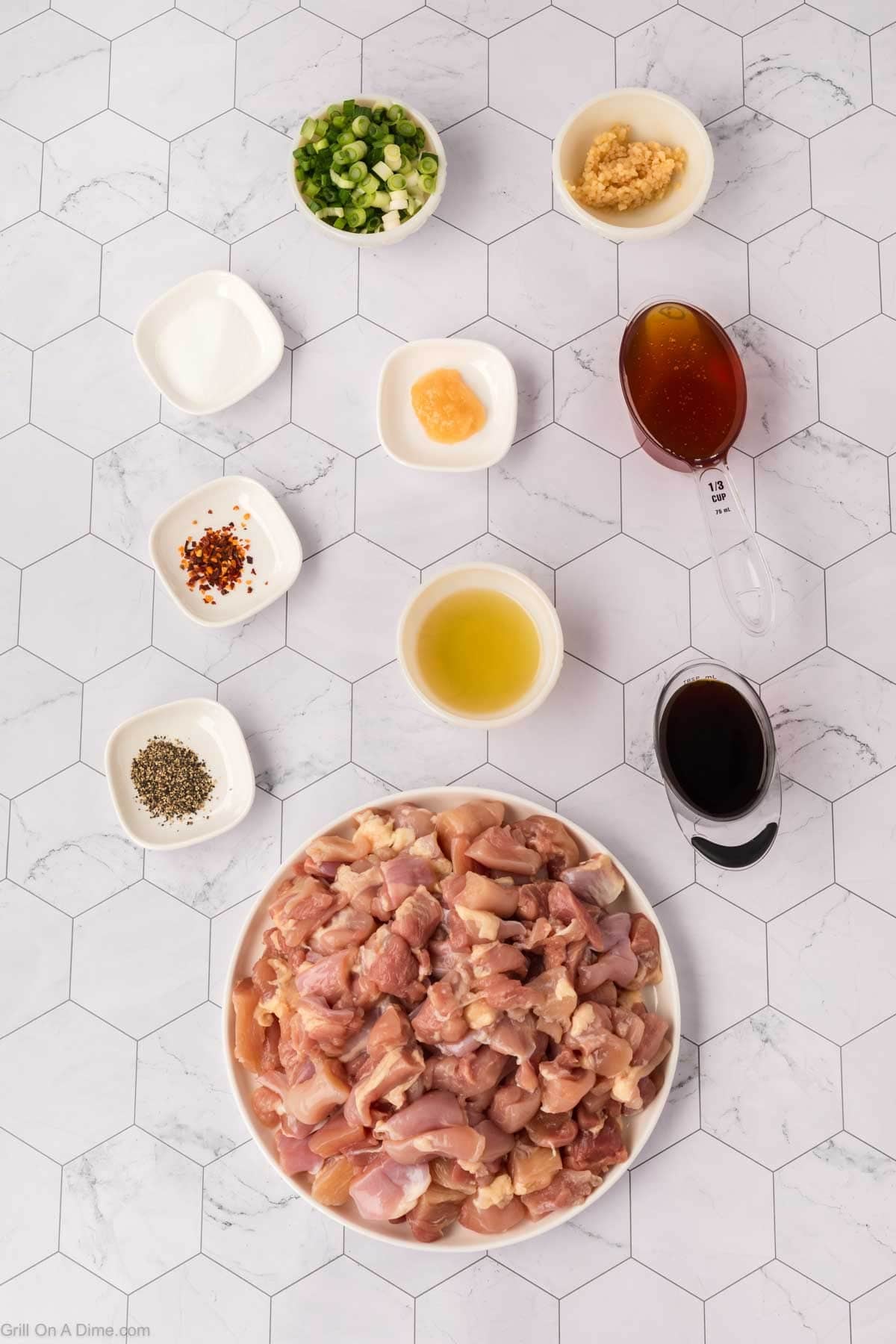 A plate of raw, chopped chicken—perfect for Blackstone Chicken—is surrounded by bowls of green onions, minced garlic, honey, soy sauce, and seasonings on a white hexagon-tiled surface, ready for a delicious Honey Garlic Chicken recipe.