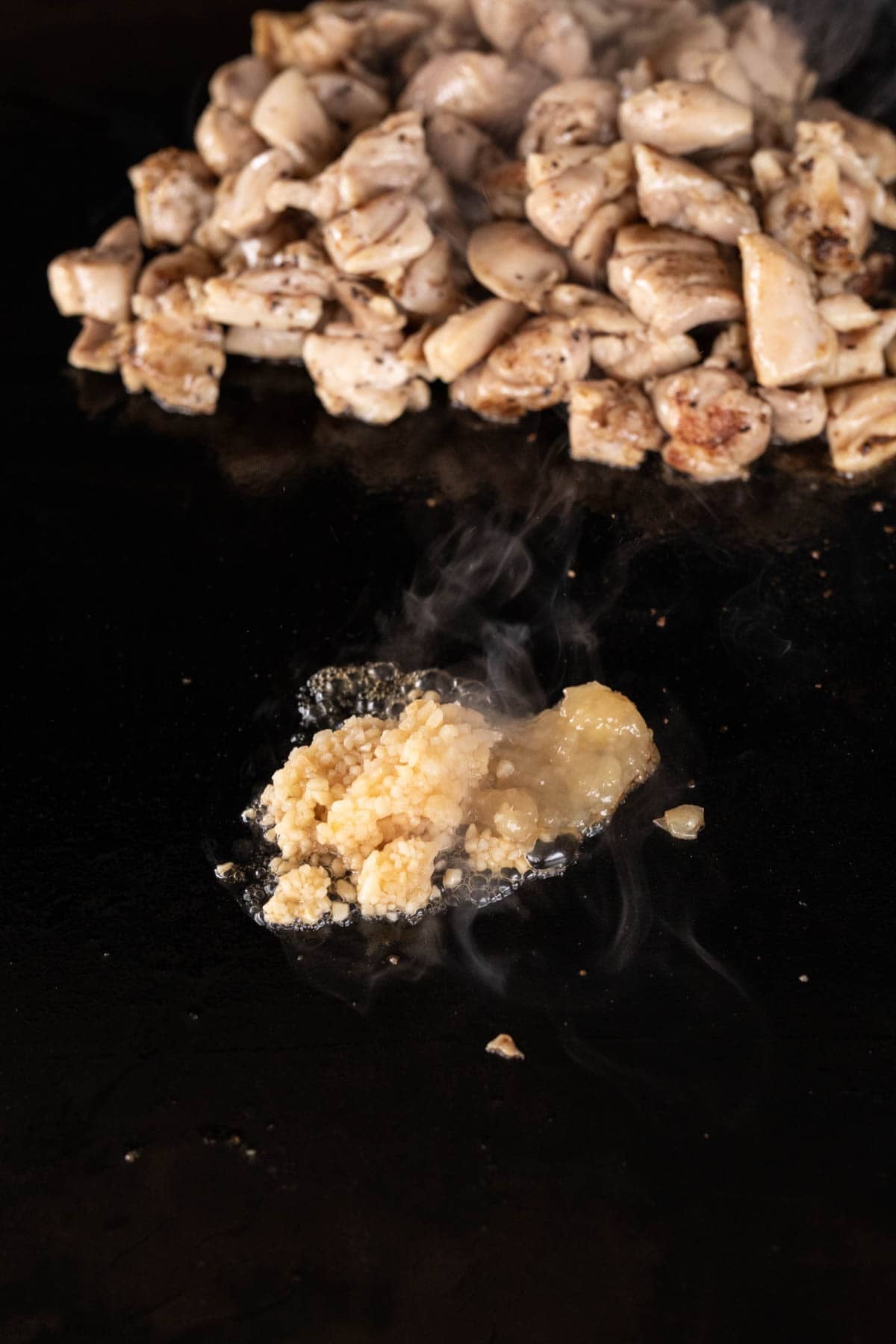 Minced garlic and ginger sizzle on a Blackstone griddle, while a pile of cooked, diced chicken breast waits nearby. Steam rises from the fragrant mix—perfect beginnings for delicious Honey Garlic Chicken.