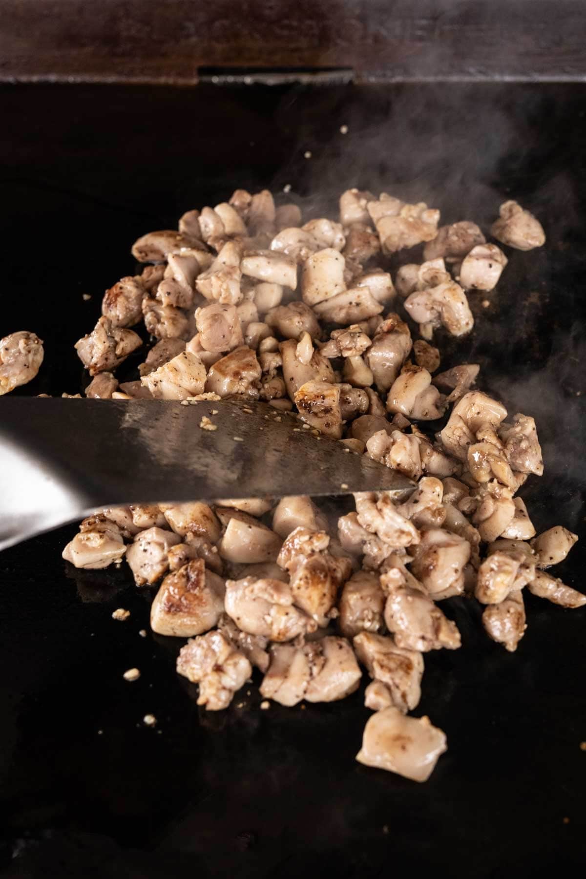 Diced pieces of chicken sizzling on a Blackstone grill, with steam rising and a metal spatula stirring the Honey Garlic Chicken for a mouthwatering finish.