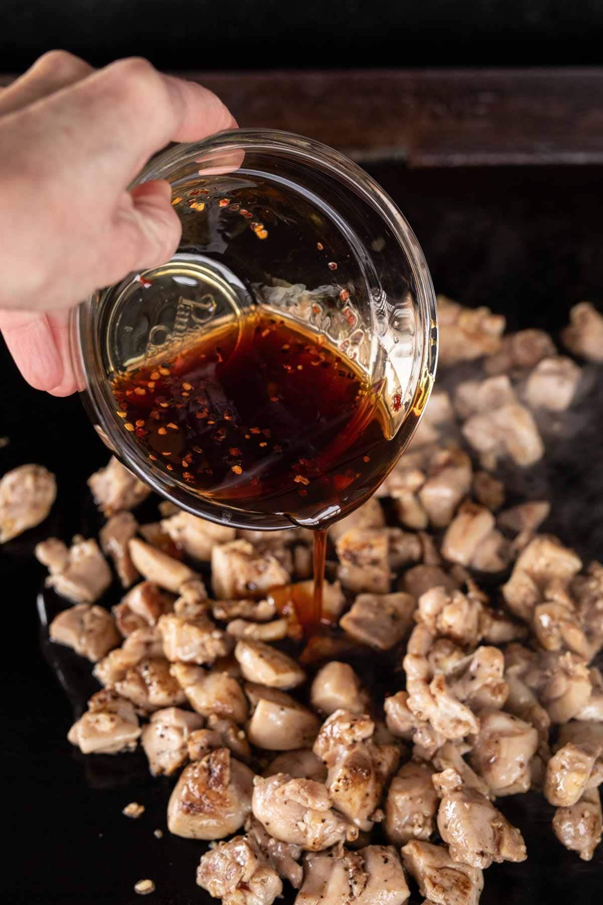 A hand pours dark, seasoned sauce from a glass bowl onto a pan filled with sizzling, diced pieces of cooked Honey Garlic Chicken. Steam rises from the hot surface, capturing this mouthwatering chicken recipe in action.