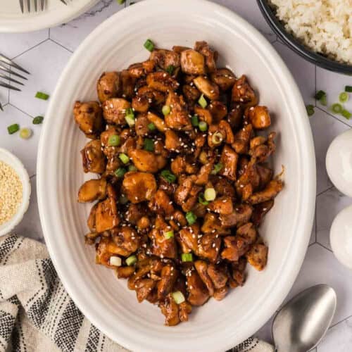 A white oval plate filled with glazed, diced Honey Garlic Chicken garnished with chopped green onions and sesame seeds, surrounded by bowls of rice and sesame seeds, a spoon, and a checkered cloth napkin—perfect for Blackstone Chicken recipes.