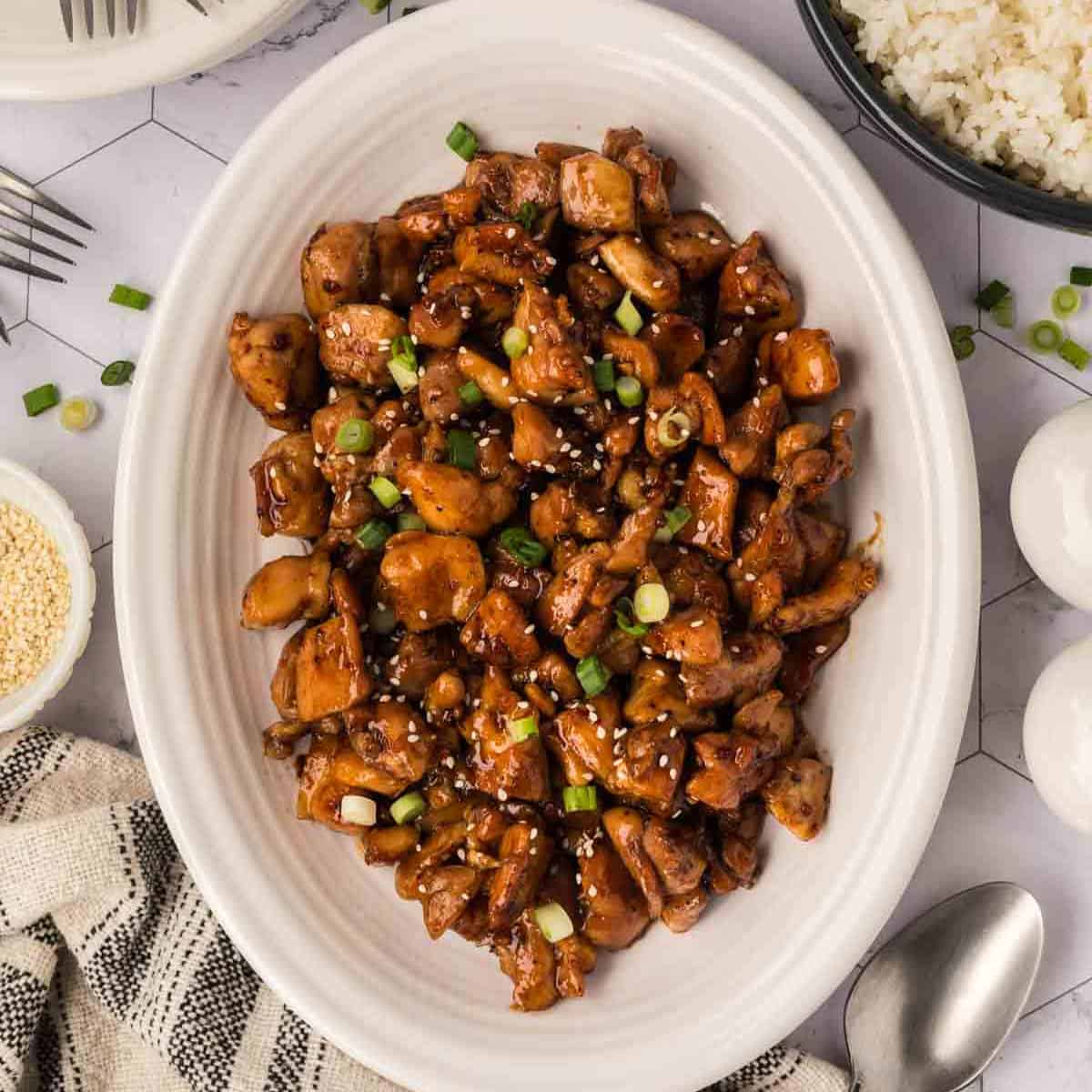 A white oval plate filled with glazed, diced Honey Garlic Chicken garnished with chopped green onions and sesame seeds, surrounded by bowls of rice and sesame seeds, a spoon, and a checkered cloth napkin—perfect for Blackstone Chicken recipes.