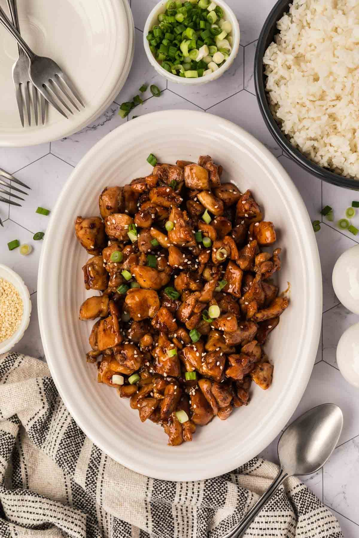 A white oval plate of Honey Garlic Chicken garnished with sesame seeds and chopped green onions. Surrounding it are bowls of rice, sliced green onions, sesame seeds, stacked plates, forks, and a striped napkin.