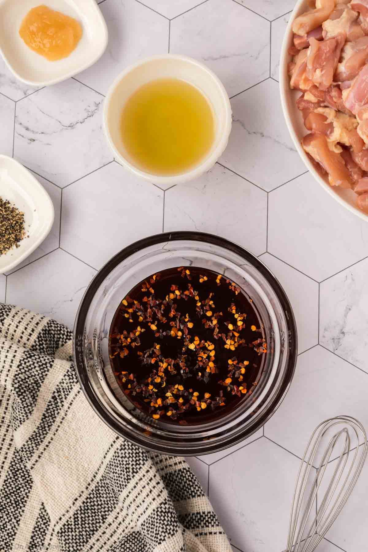 A bowl of dark marinade with red pepper flakes sits on a hexagon-tiled counter, ready for making Honey Garlic Chicken, surrounded by raw chicken pieces, olive oil, black pepper, honey, a whisk, and a black-and-white plaid towel.