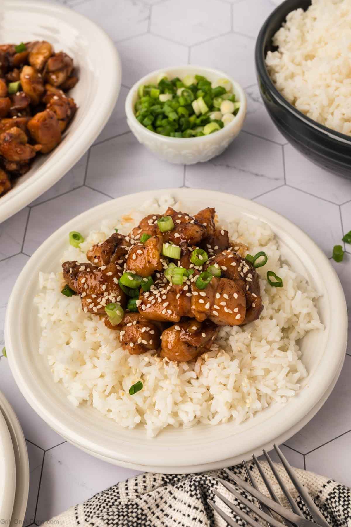A plate of white rice topped with Honey Garlic Chicken, sprinkled with sesame seeds and chopped green onions. In the background, a bowl of rice, more glazed Blackstone Chicken, and green onions complete this delicious Blackstone Recipe.