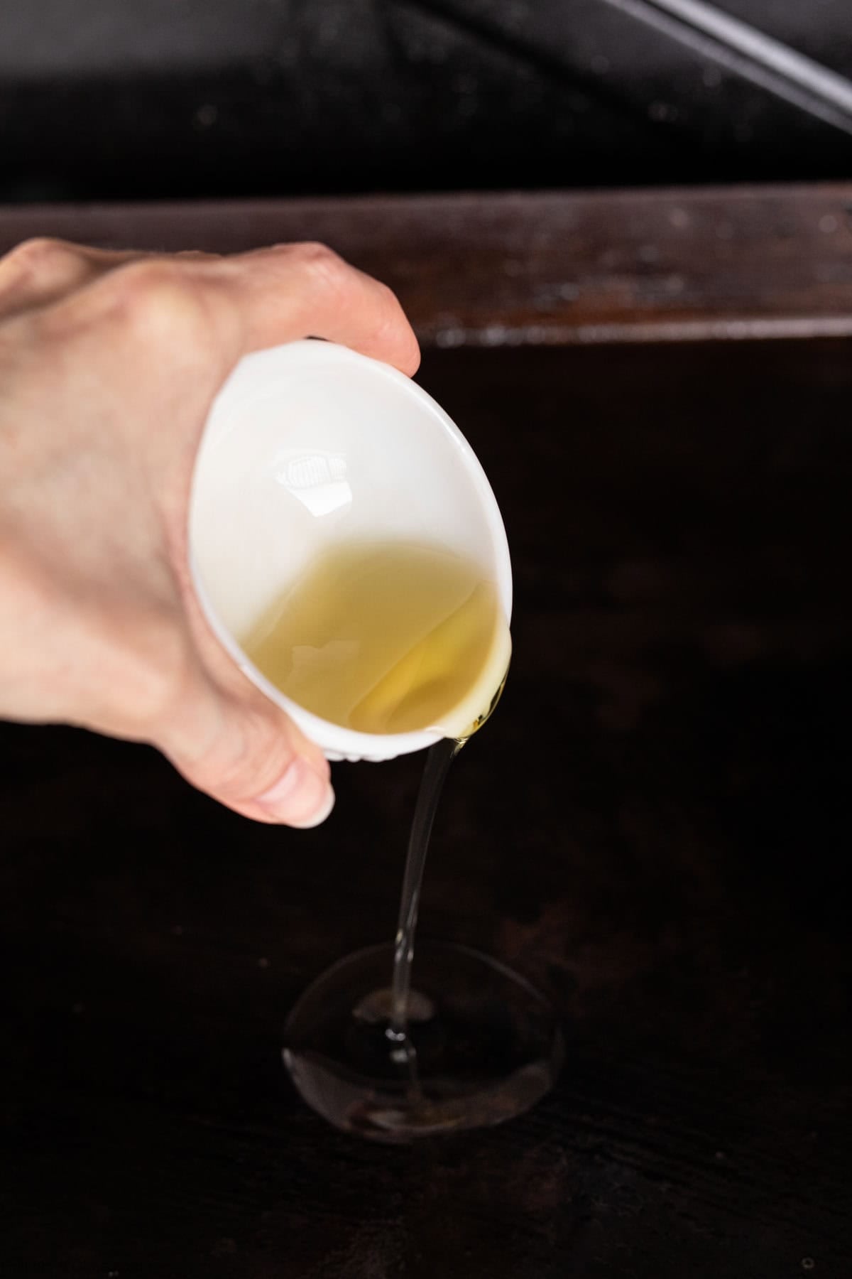 A hand pours a light yellow liquid, possibly oil for Honey Garlic Chicken, from a small white cup onto a dark surface—perfect for starting your favorite Blackstone Recipes.