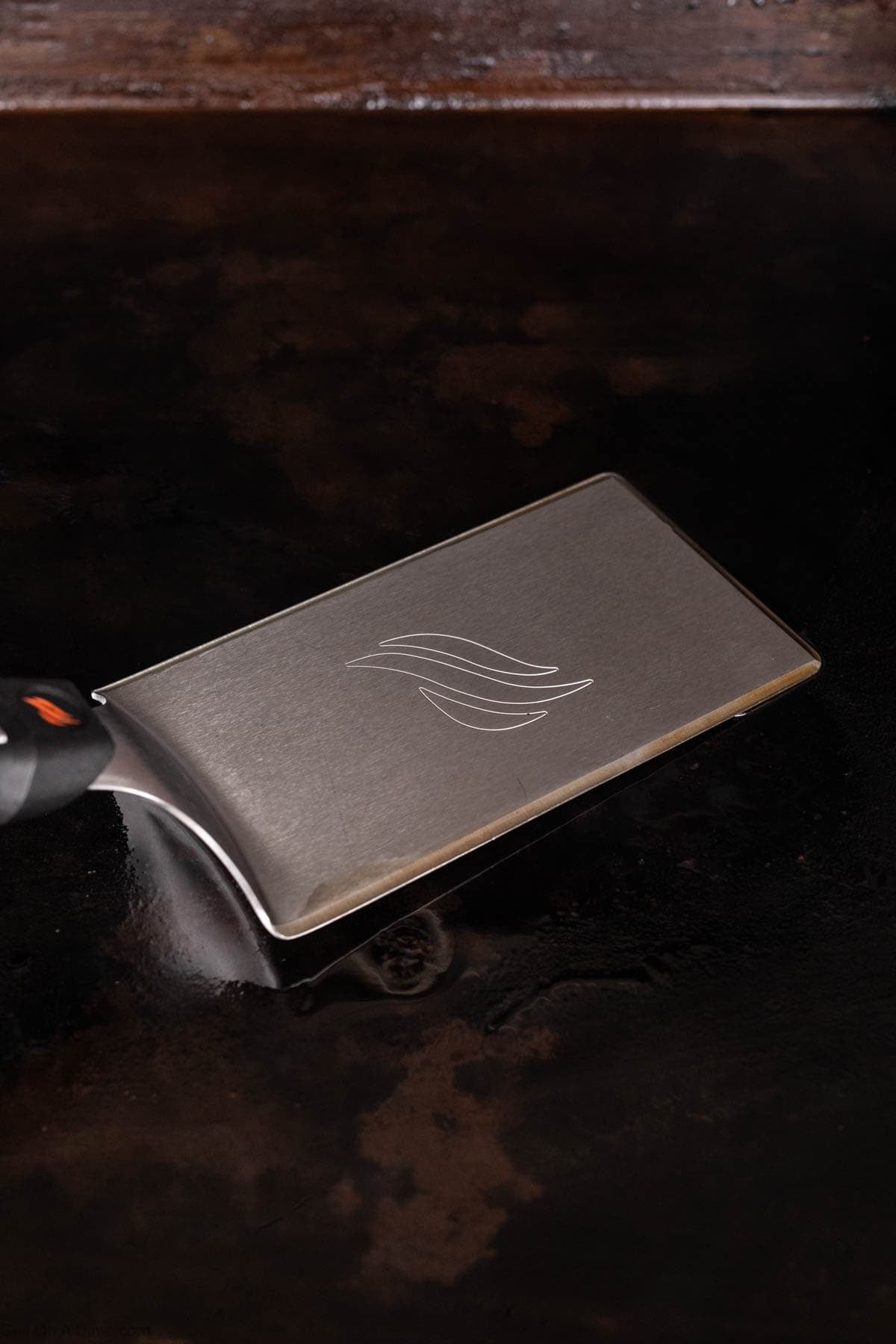 A metal spatula with a black handle rests on a dark, flat surface. Perfect for flipping Blackstone Chicken, the spatula’s blade features a simple wave-like design etched on it.