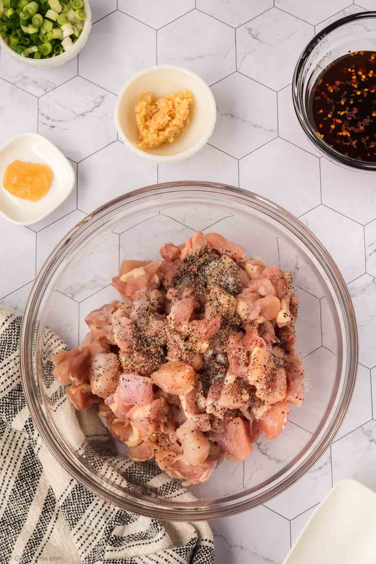 A glass bowl filled with raw, diced chicken seasoned with black pepper sits on a countertop, ready for a delicious Honey Garlic Chicken—perfect for trying out new Blackstone Recipes—surrounded by small bowls of green onions, garlic, ginger, and sauce.