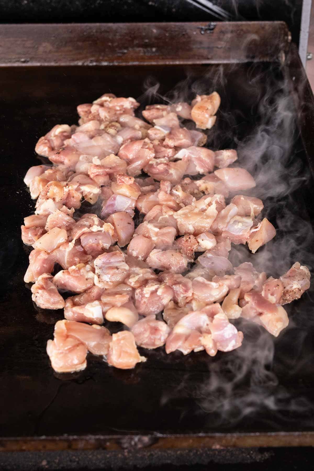 Chopped raw chicken pieces cooking on a hot, black griddle with steam rising around them—a perfect start to a flavorful Blackstone Chicken recipe.