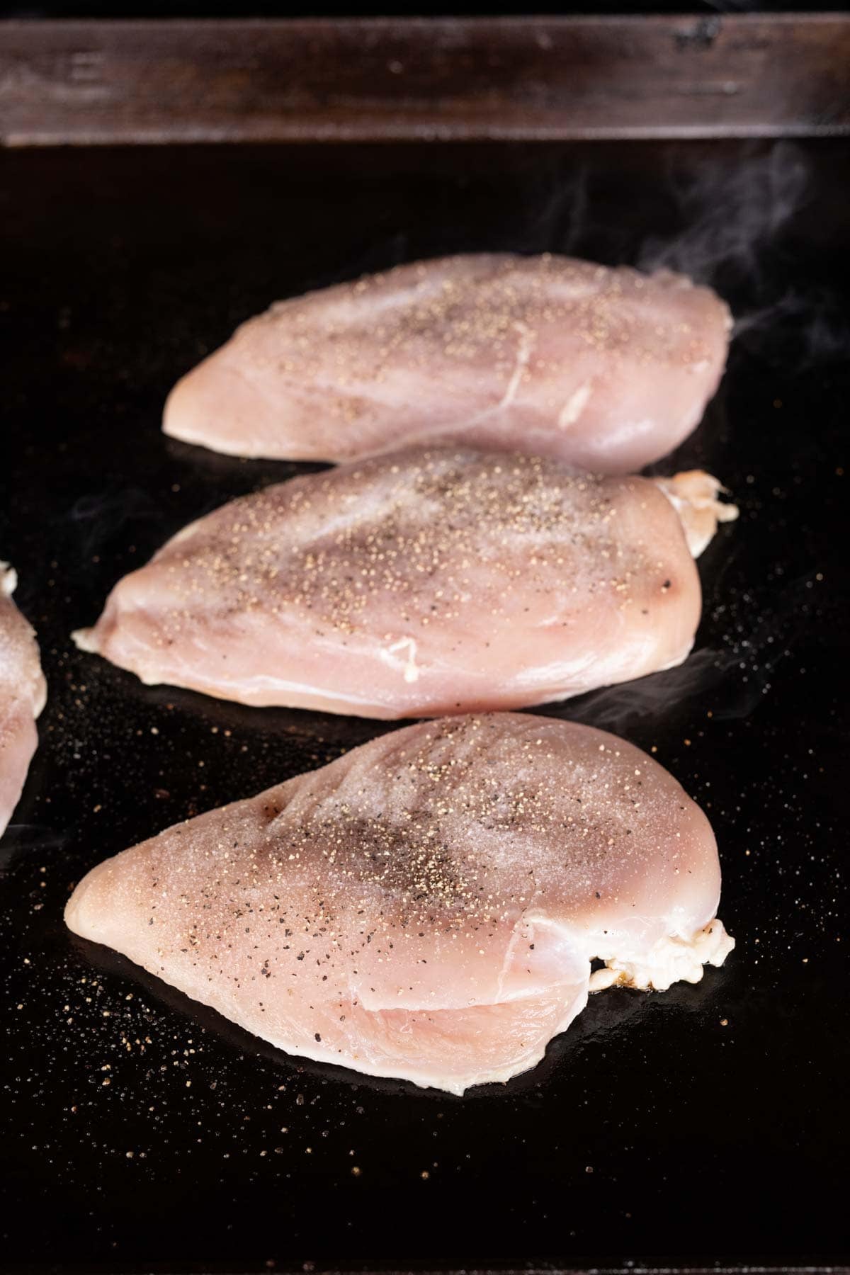 Three raw chicken breasts, seasoned with salt and pepper, sit on a Blackstone griddle ready to become a flavorful Loaded Chicken dish. Steam or smoke rises around the edges, promising a tasty Blackstone recipe in the making.