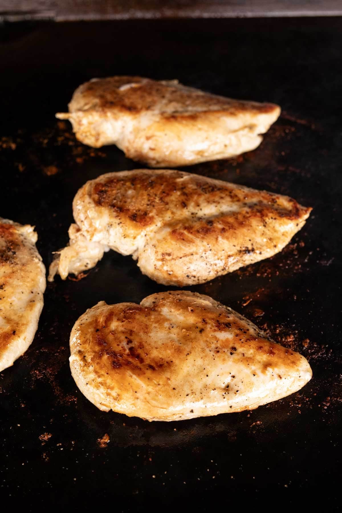 Four seasoned chicken breasts sizzle on a dark Blackstone griddle. This loaded chicken is golden brown with visible spices, and the surface around them appears slightly oily and delightfully charred.