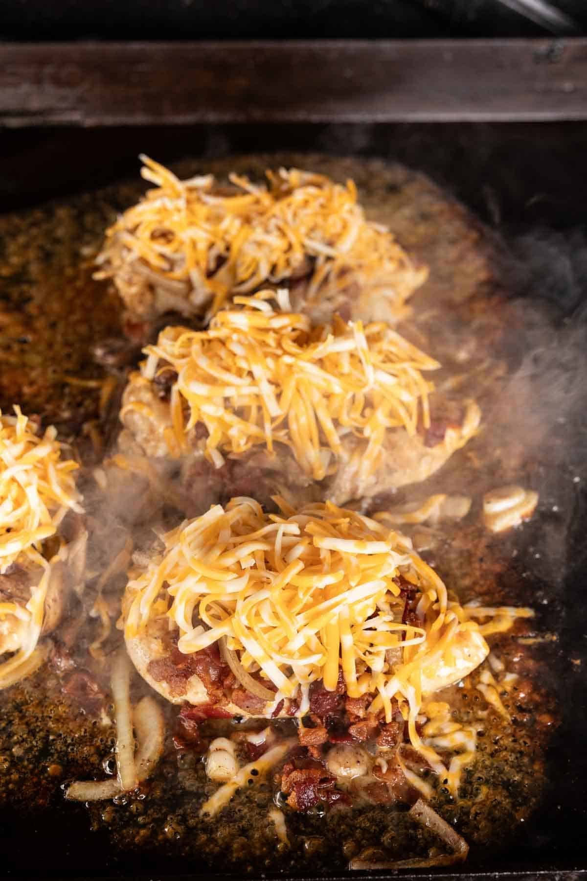 Three cooked chicken breasts on a skillet, topped with a generous amount of shredded yellow and white cheese, onions, and steam rising from the hot surface—a delicious Loaded Chicken dish perfect for your Blackstone griddle.