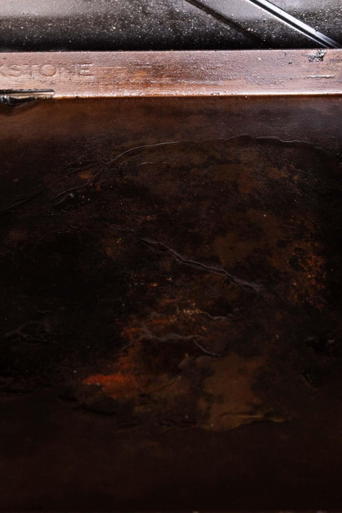 A close-up of a dark, rusted, and weathered metal surface with some scratches and brownish-red patches, bordered by a slightly lighter brown metallic edge—reminiscent of a well-used Blackstone griddle.