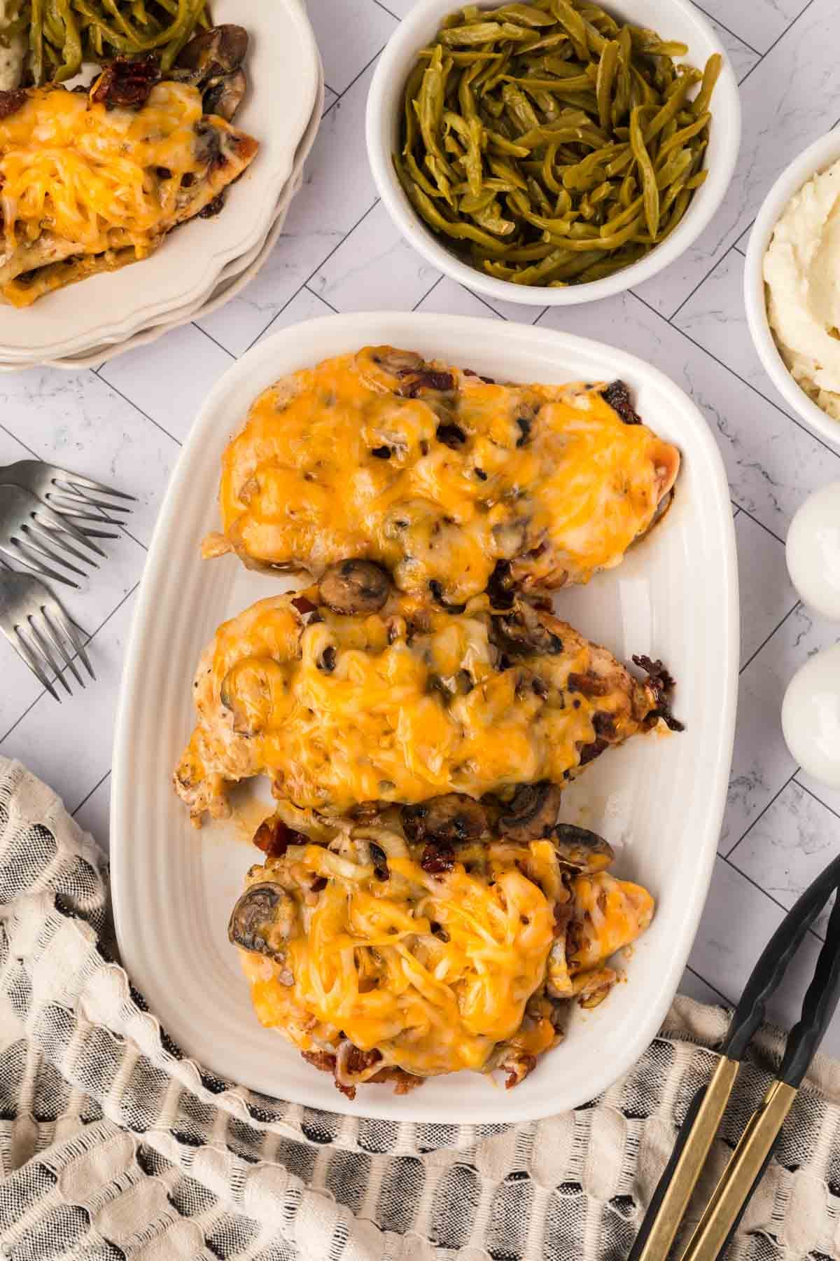 A serving platter with three Loaded Chicken breasts topped with melted cheese, mushrooms, and bacon, surrounded by bowls of green beans and mashed potatoes on a tiled table—perfect for your next Blackstone cookout.