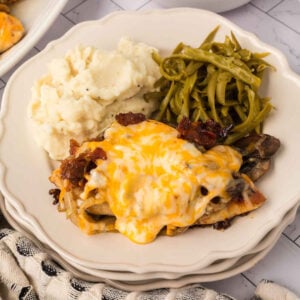 A plate with mashed potatoes, green beans, and a serving of Loaded Chicken topped with melted cheese and bacon pieces.