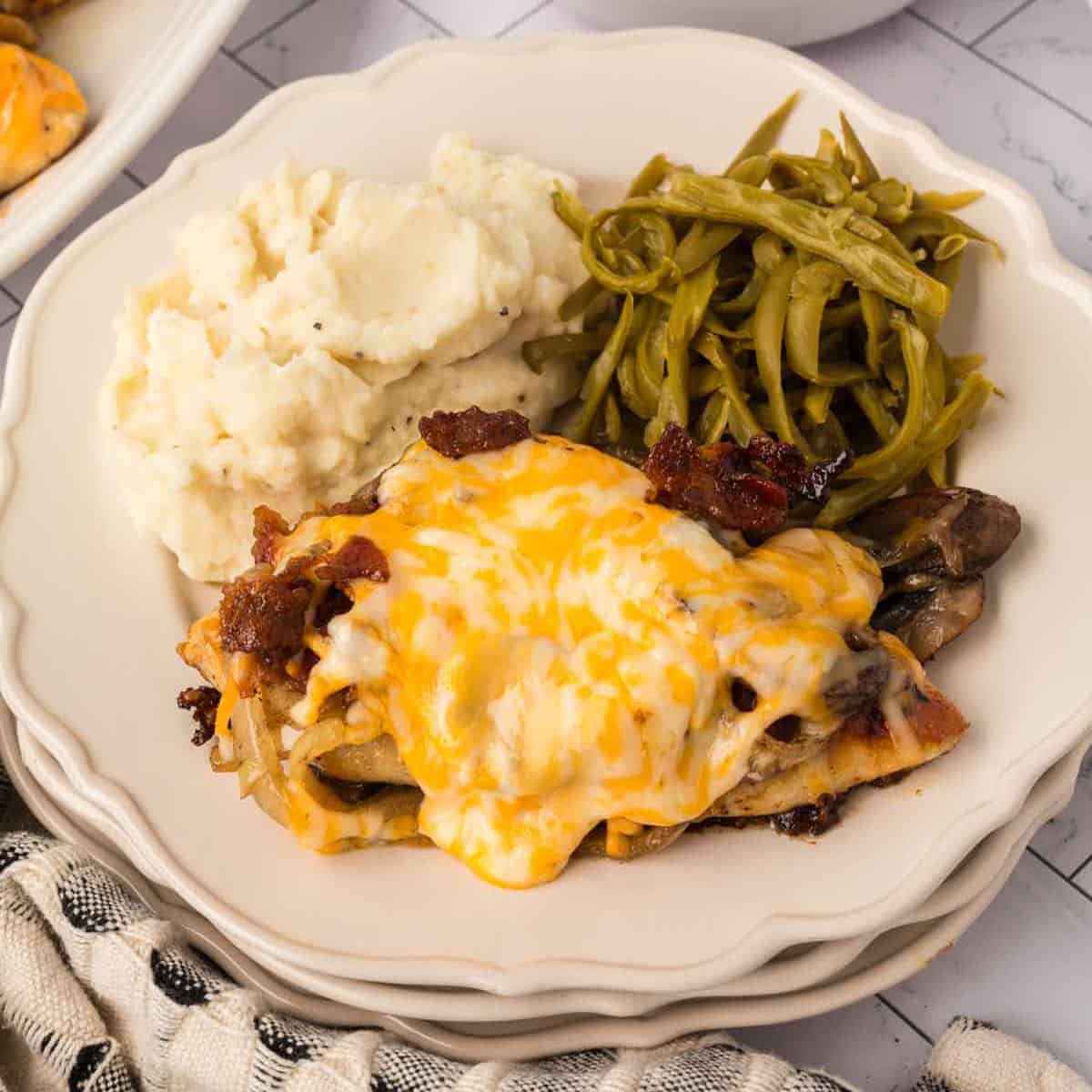 A plate with mashed potatoes, green beans, and a serving of Loaded Chicken topped with melted cheese and bacon pieces.