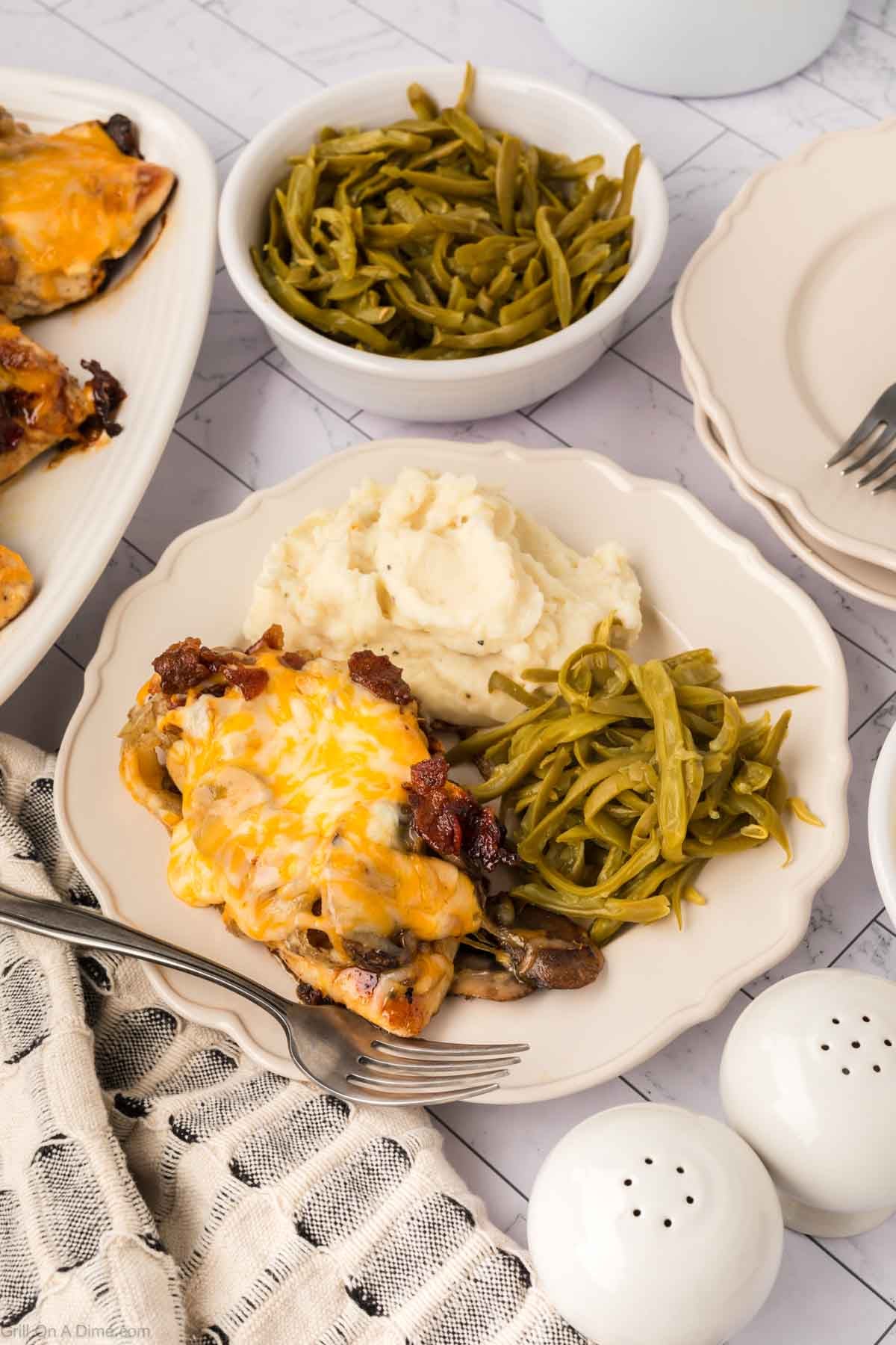 A plate of food with mashed potatoes, green beans, and cheesy Blackstone Chicken sits next to a fork and napkin. Nearby are bowls of green beans, a stack of plates, and salt and pepper shakers.