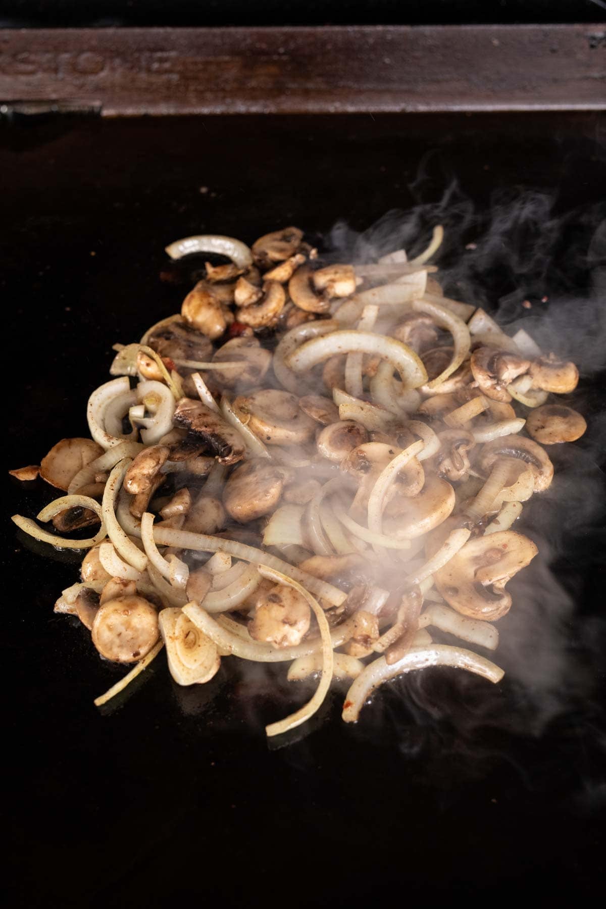 Sliced onions and mushrooms sizzling on a hot Blackstone griddle, with visible steam rising from the vegetables as they cook.