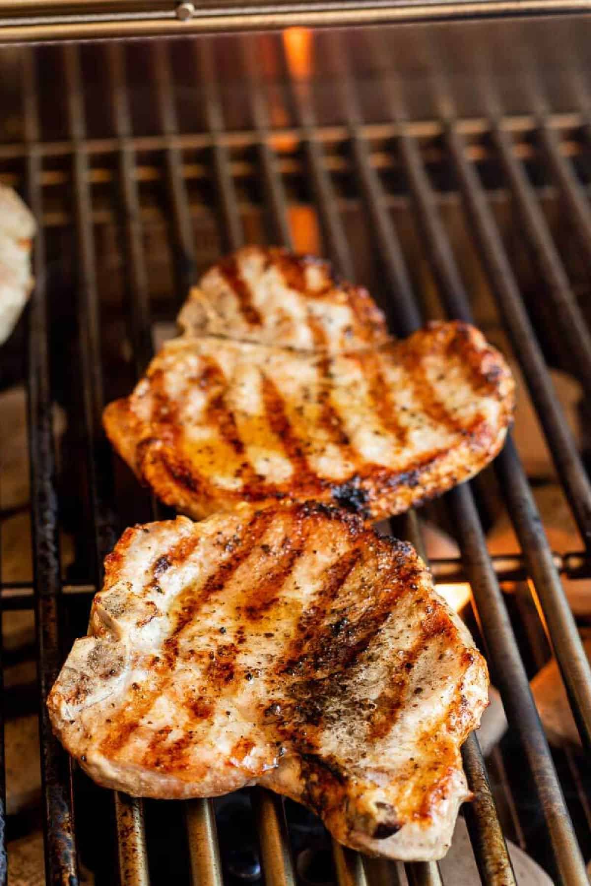 Two Grilled BBQ Bone-In Pork Chops with perfect grill marks sizzle over open flames on a barbecue, the smoky aroma rising from the hot metal grates.