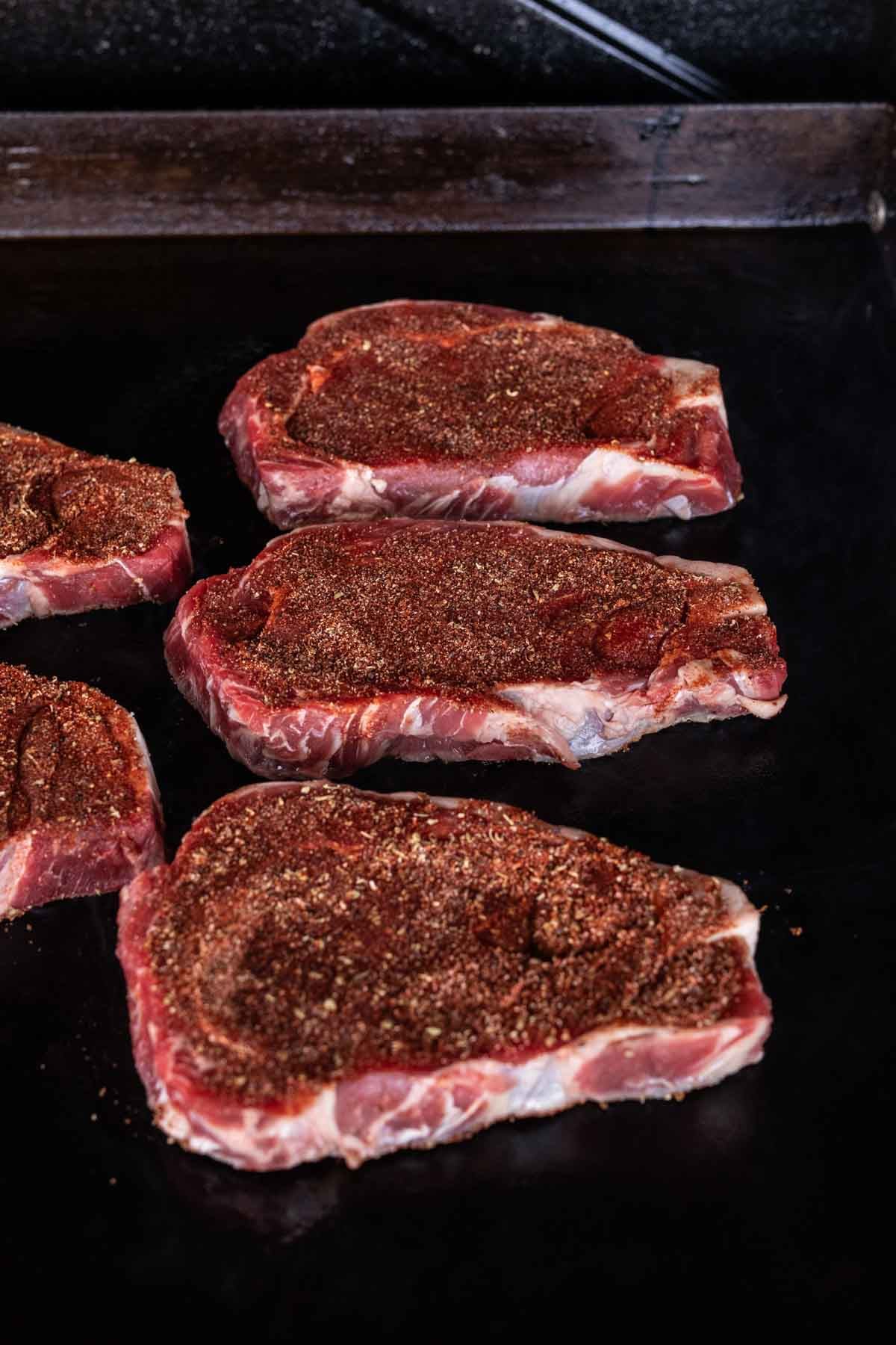 Four raw, seasoned Blackened Steaks rest on a dark Blackstone griddle, ready to be cooked. The rich seasoning coats each steak, giving them a bold, reddish-brown appearance.