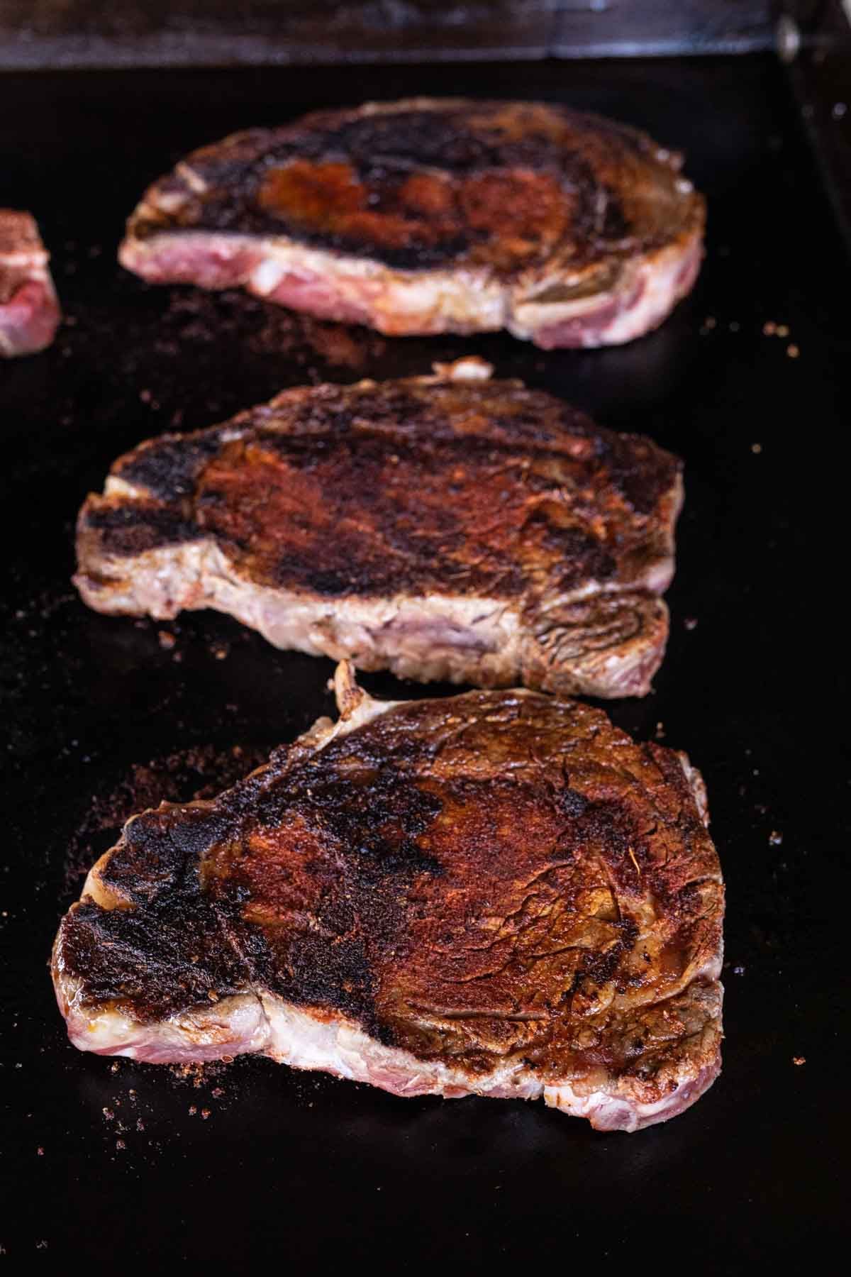Three seared ribeye steaks with a dark, crispy crust sizzle on a flat Blackstone griddle. The Blackened Steaks are spaced apart and appear juicy and well-seasoned.