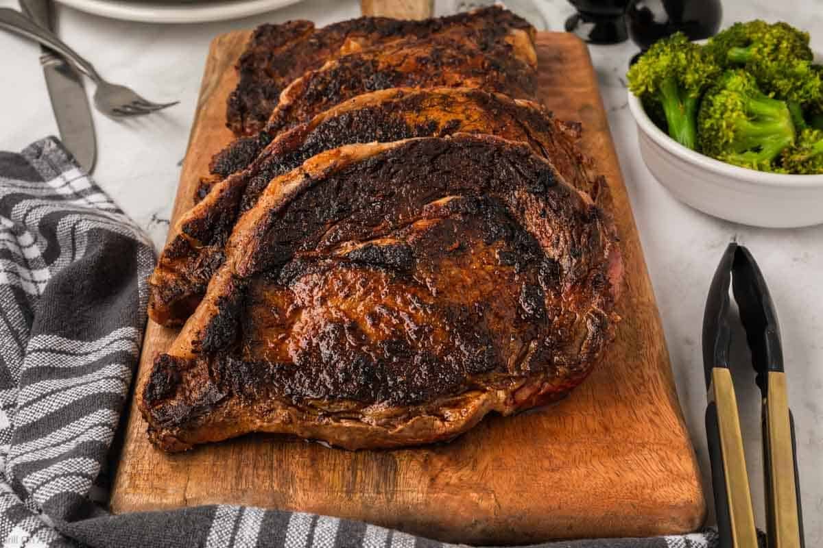 Several slices of perfectly grilled blackened steak are arranged on a wooden cutting board, with a striped towel, fork, and a bowl of broccoli nearby on a light-colored surface—an inviting Blackstone steak recipe scene.