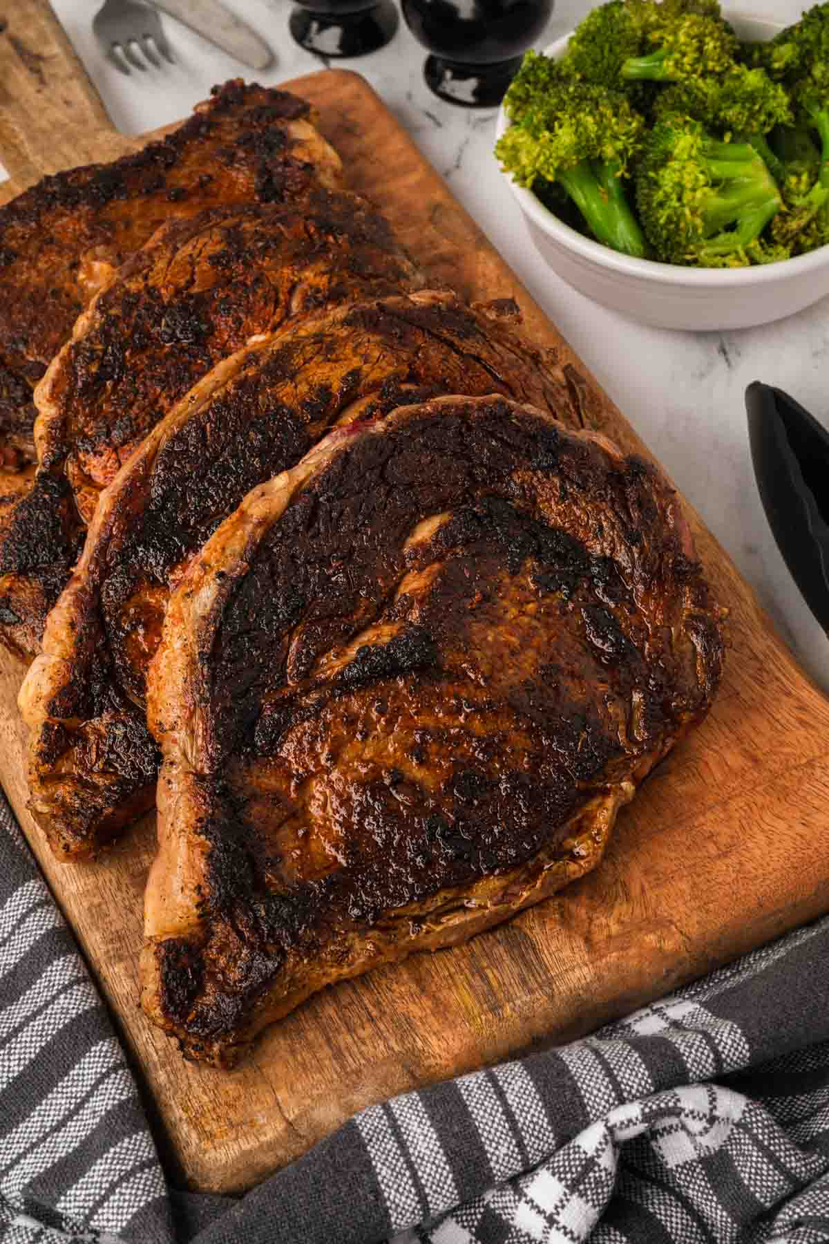 Four grilled Blackened Steaks with a charred crust are arranged on a wooden cutting board, next to a bowl of steamed broccoli and a black-and-white checkered cloth.