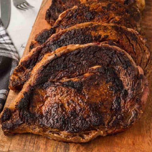Four Blackened Steaks with a dark, charred crust are arranged on a wooden cutting board, fresh from the Blackstone. A striped towel and fork are partially visible in the background.