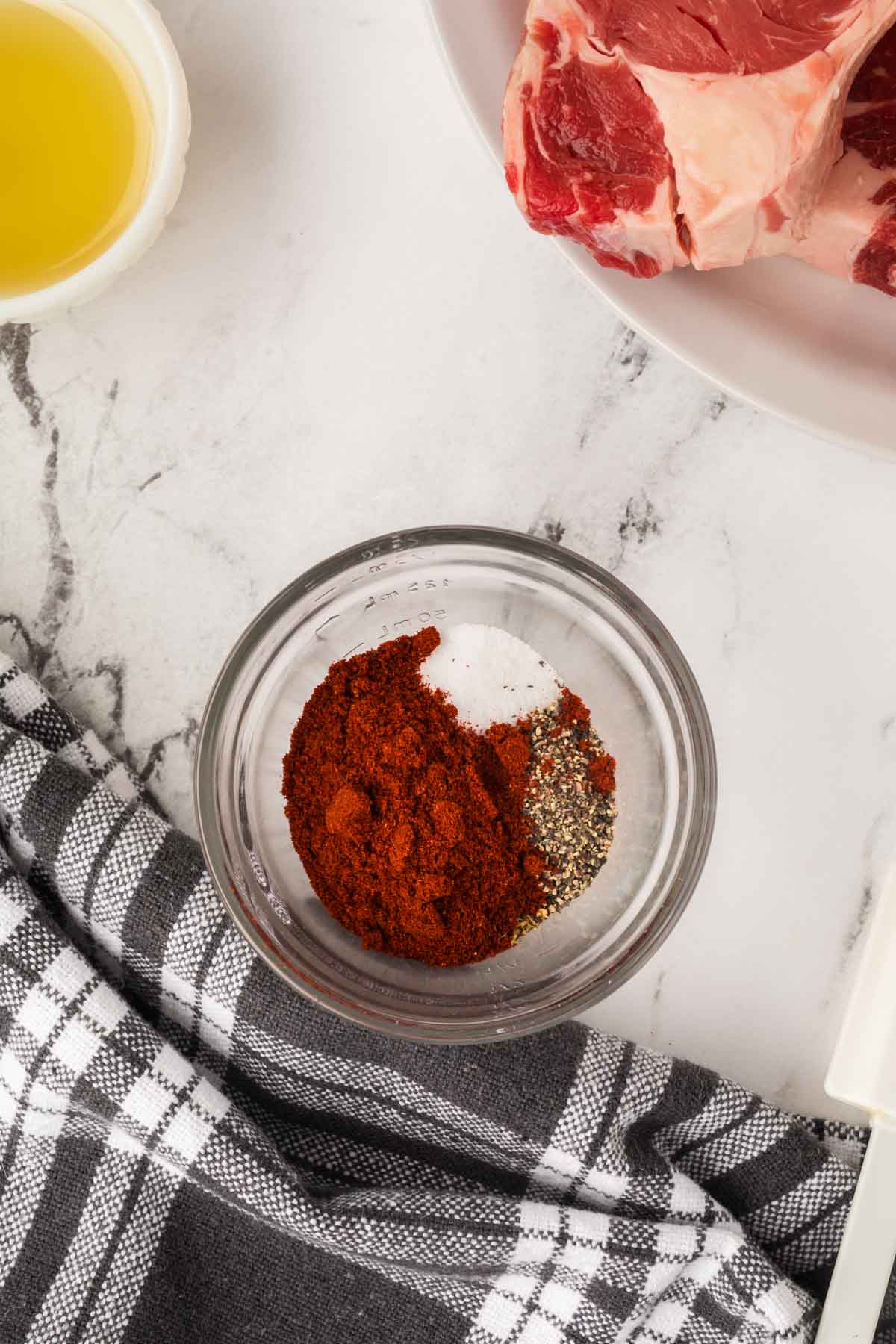 A small glass bowl with paprika, salt, and black pepper sits on a marble surface, next to a checkered cloth, olive oil, and a plate of raw steak—perfect for seasoning your Blackened Steak before grilling on the Blackstone.