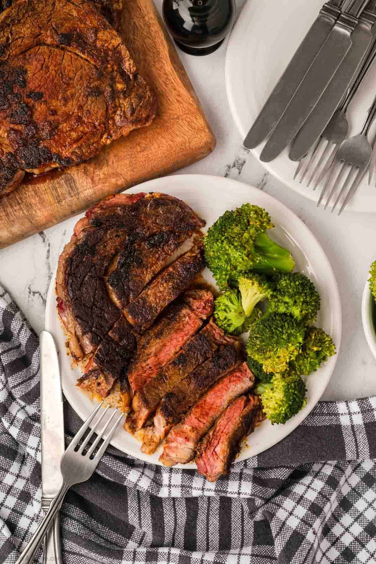A plate with sliced Blackened Steak and steamed broccoli sits next to a fork and knife. A larger Blackstone-grilled steak rests on a wooden board, with utensils and a black-and-white checkered napkin nearby.