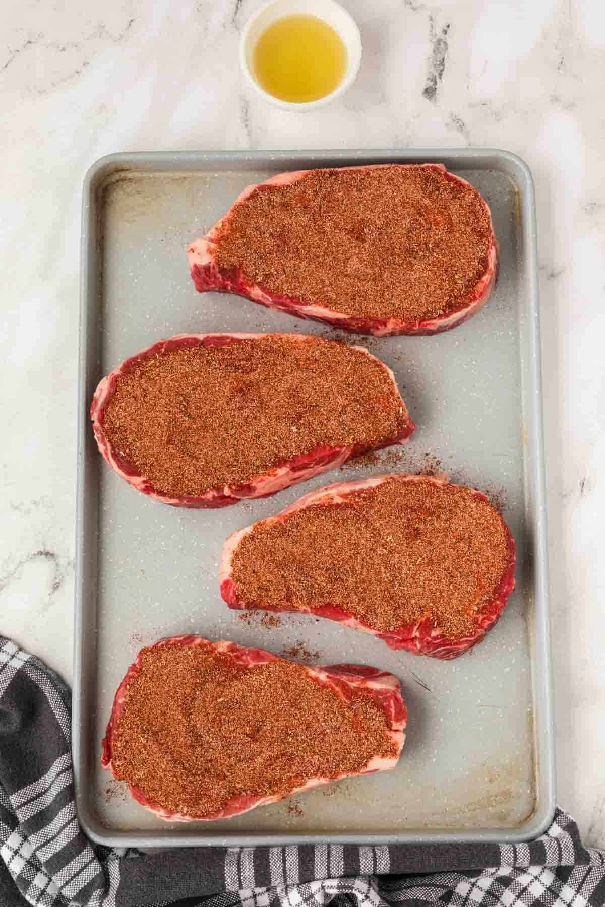 Four raw Blackened Steaks, coated with a flavorful dry spice rub, are arranged on a gray baking sheet. A small bowl of oil sits above the sheet, and a black-and-white kitchen towel is partially visible in the corner.
