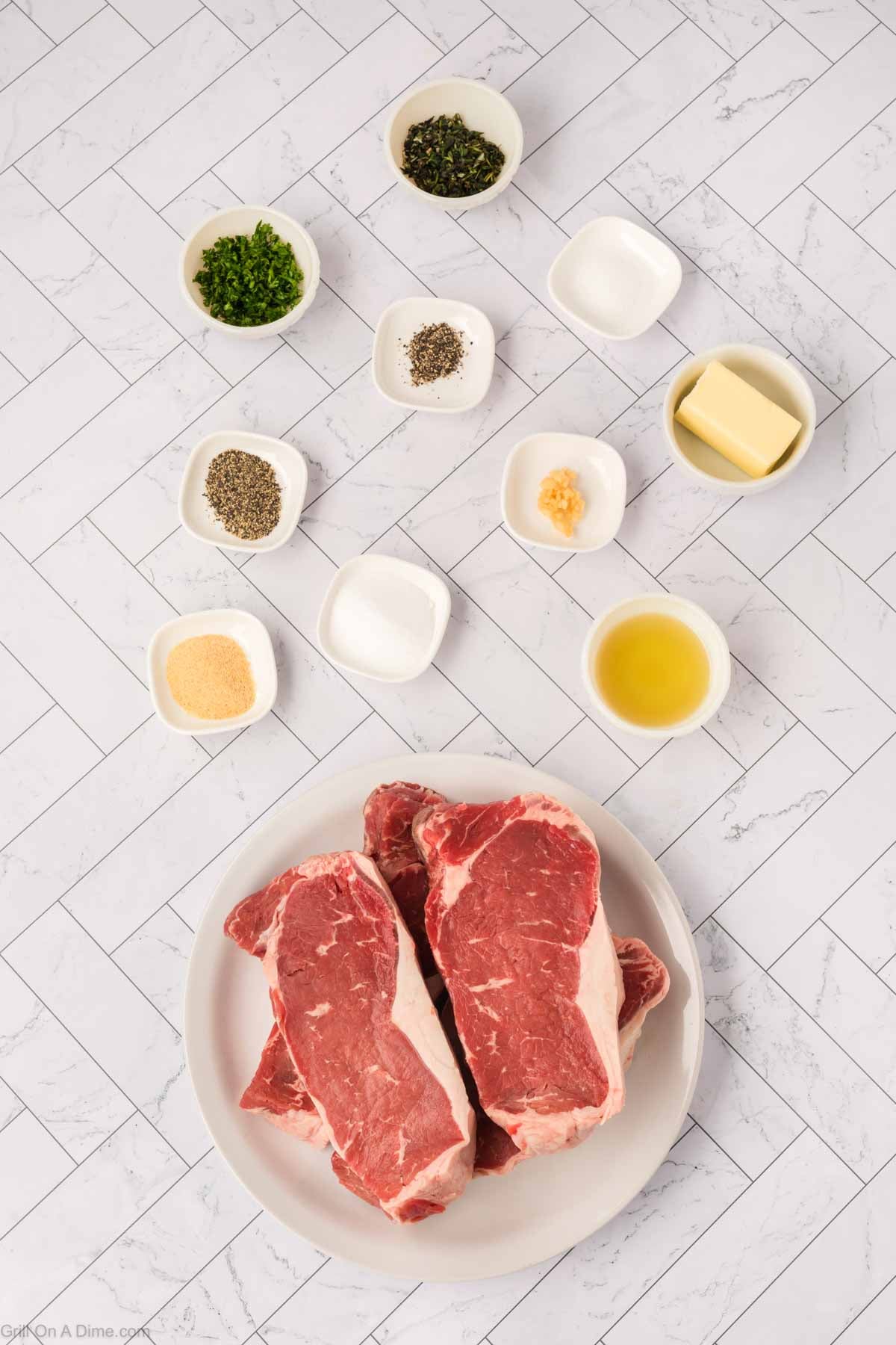 Overhead view of two raw New York Strip steaks on a plate, surrounded by small bowls of herbs, spices, minced garlic, butter, olive oil, and seasonings on a white tiled surface—perfect for your next Blackstone cookout.