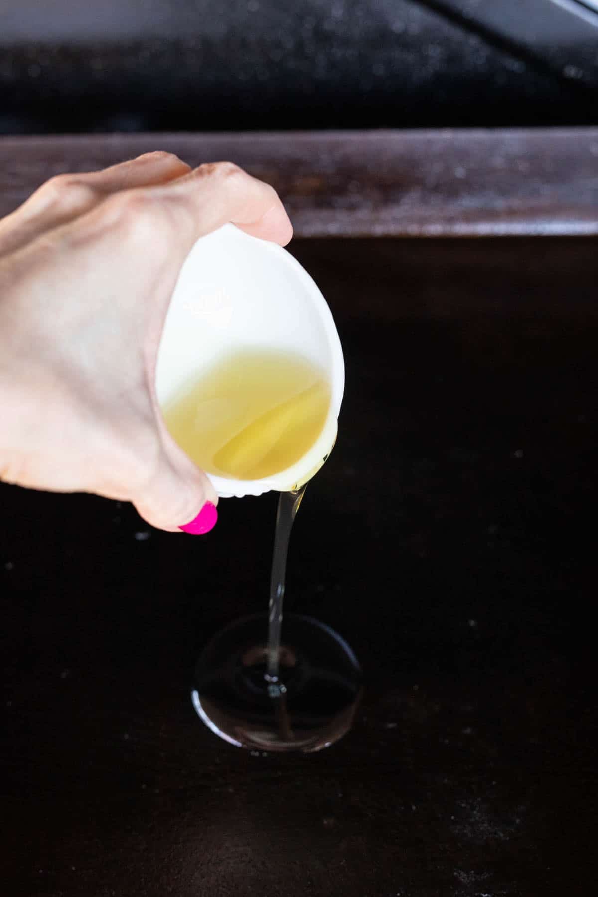 A hand pours oil from a small white cup onto a dark, flat Blackstone cooking surface, preparing it for a sizzling New York Strip.