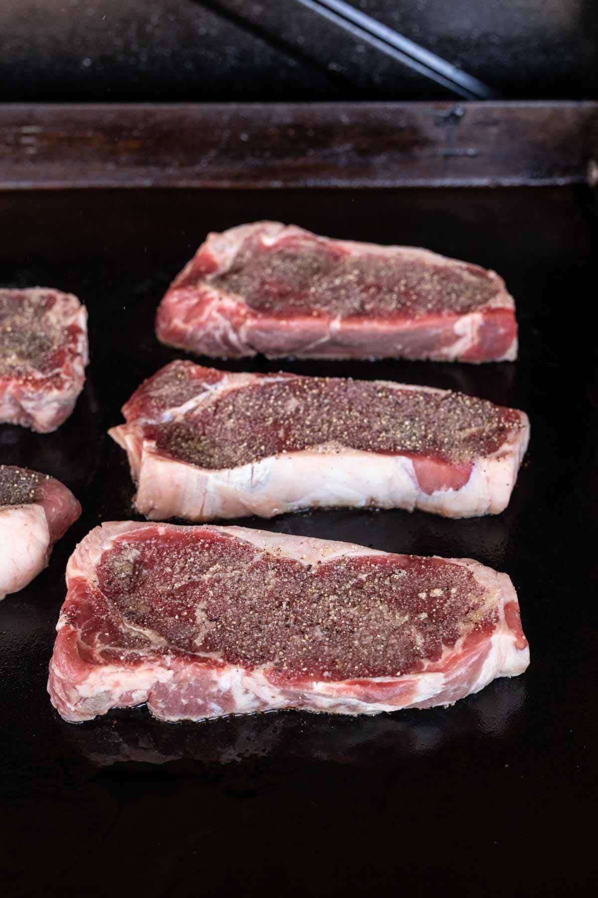 Three raw, seasoned New York Strip steaks sizzle on a Blackstone flat-top grill. The thick-cut steaks have visible fat along the edges and are generously sprinkled with black pepper and savory seasoning.