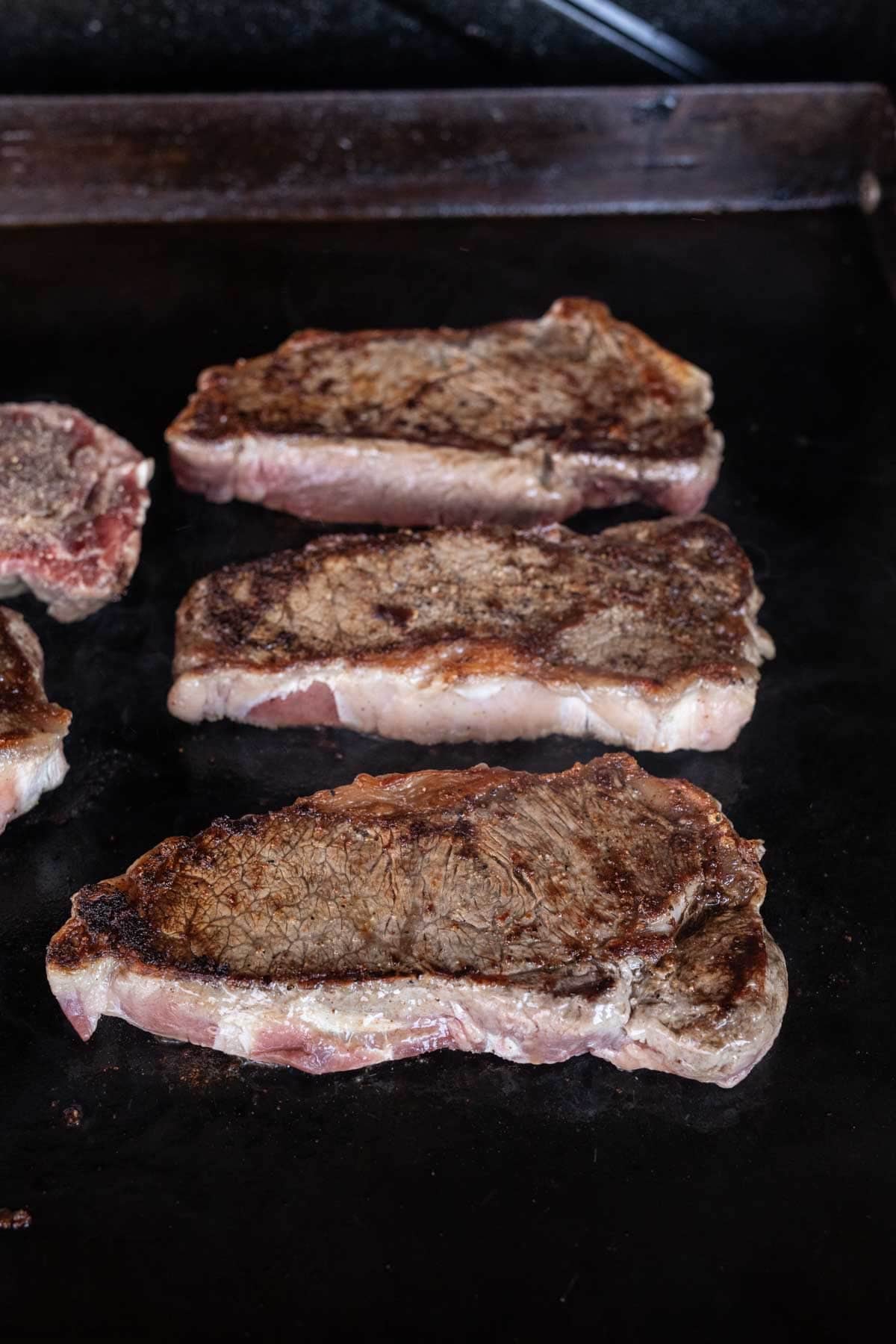 Four pieces of New York Strip steak sizzle on a dark Blackstone griddle, each boasting a beautifully browned, seared crust on the surface.