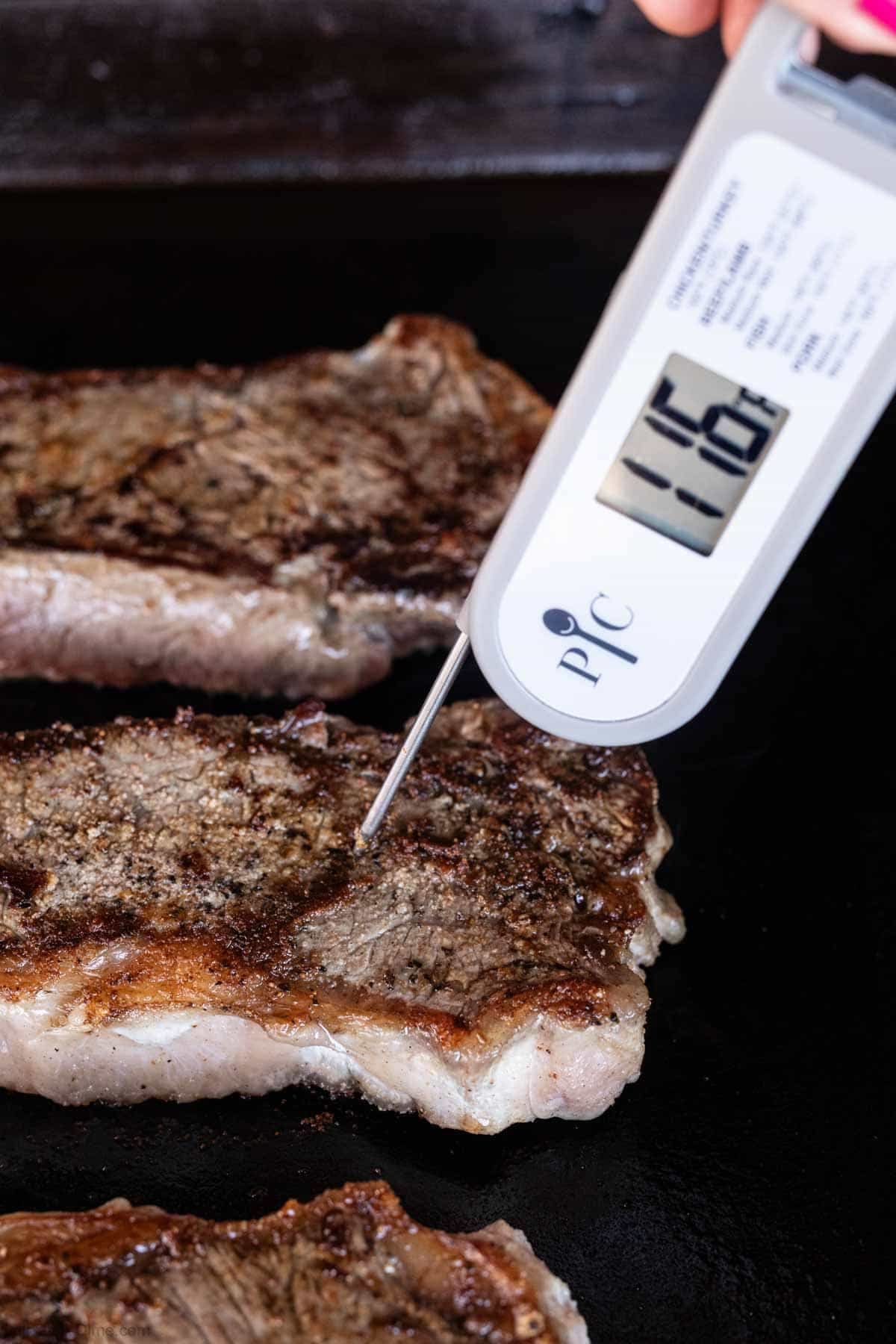 A digital meat thermometer inserted into a cooked New York Strip shows a temperature of 116°F, with two more steaks sizzling nearby on a Blackstone griddle.