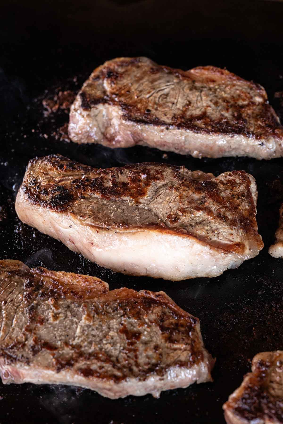 Three seared New York Strip steak pieces with a browned crust and visible fat edges cooking on a dark Blackstone grill or skillet.